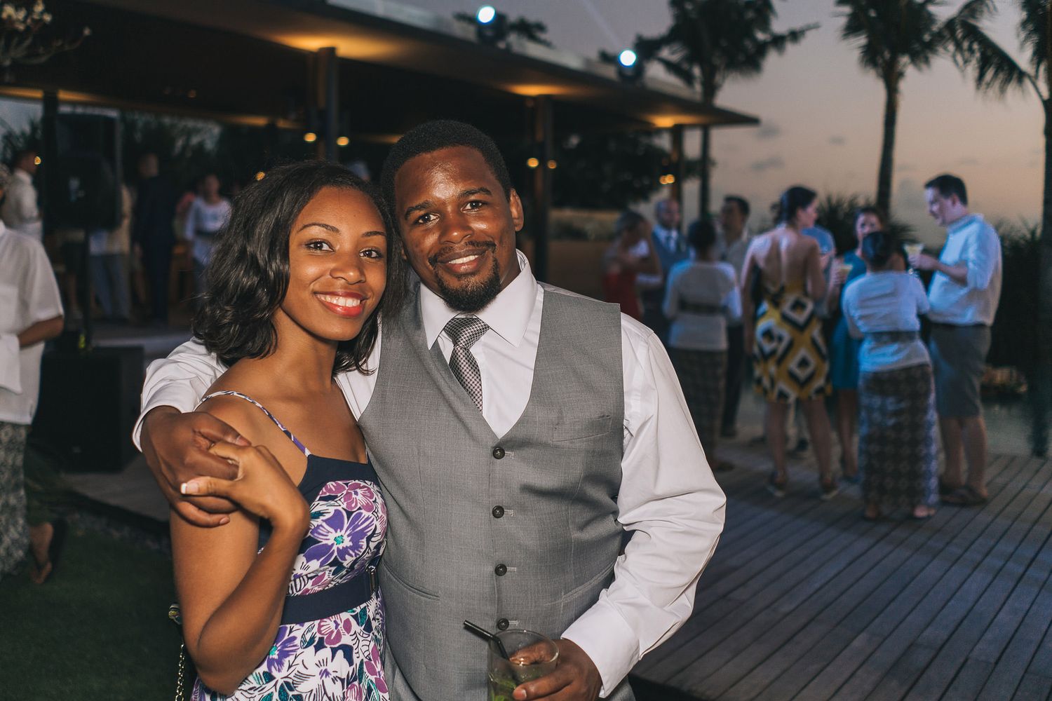 A couple enjoys an evening gathering on a wooden deck with palm trees and other guests visible in the background.