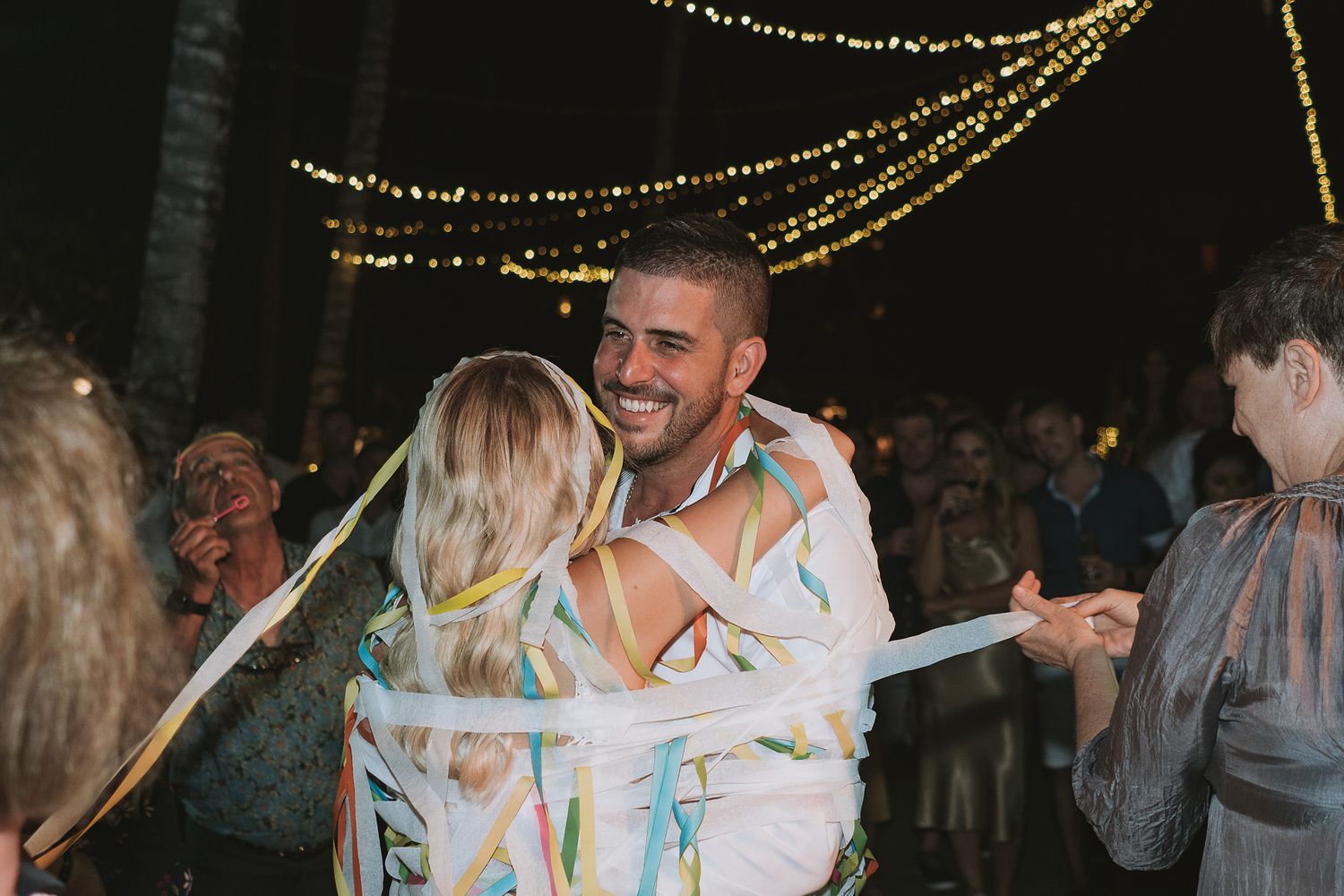 A couple dances together underneath twinkling string lights during an outdoor evening celebration.