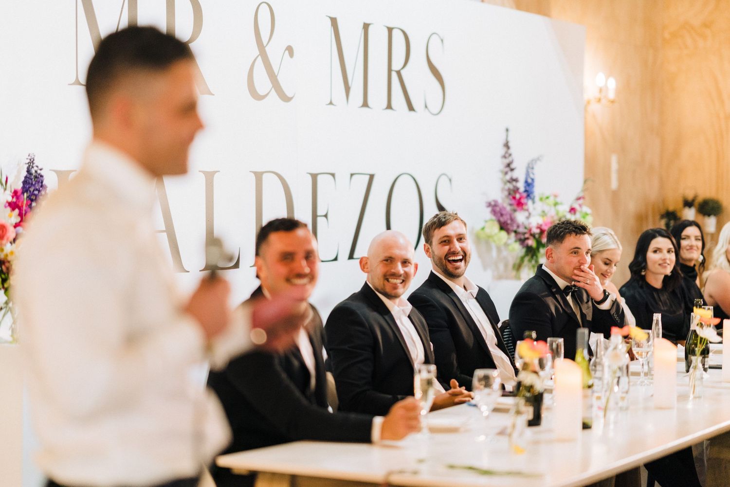 Wedding reception guests seated at a long table under a Mr & Mrs sign with floral decorations.