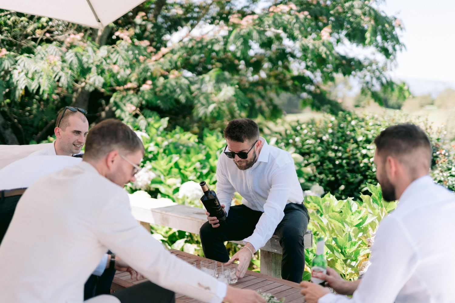 Friends gathered around a rustic outdoor dining table enjoy conversation and drinks in a lush garden setting.