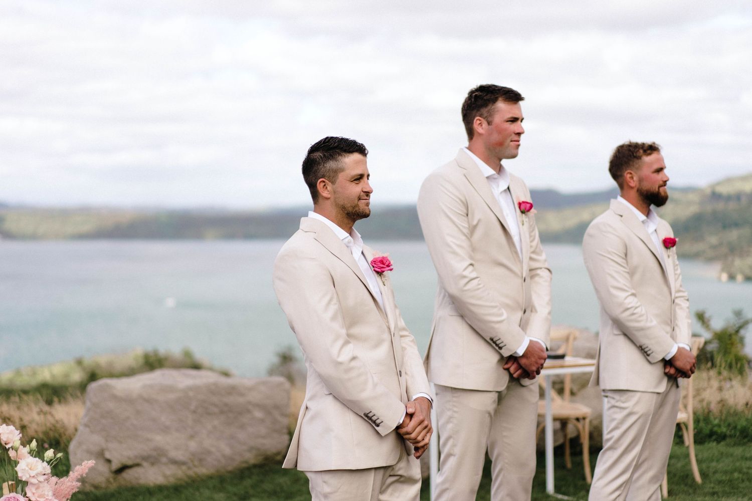 Three groomsmen in light colored suits standing at outdoor ceremony with ocean view.