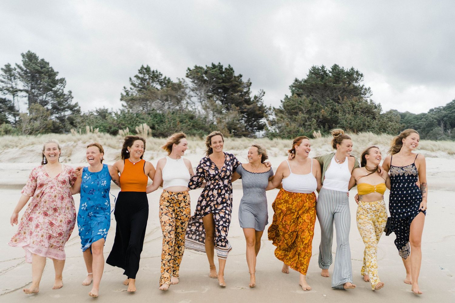 A group of women walk together down a beach wearing colorful summer dresses and jumpsuits with pine trees in background.
