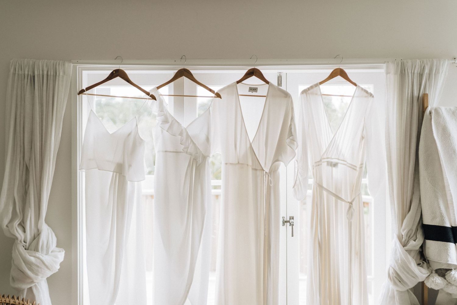 Four elegant white wedding dresses hanging on wooden hangers against a light curtained window.
