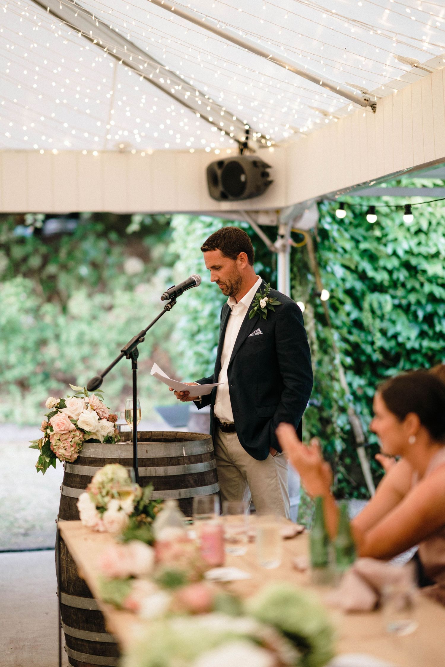 During a wedding reception, a speaker delivers a speech from behind a wooden barrel under a tent with string lights.