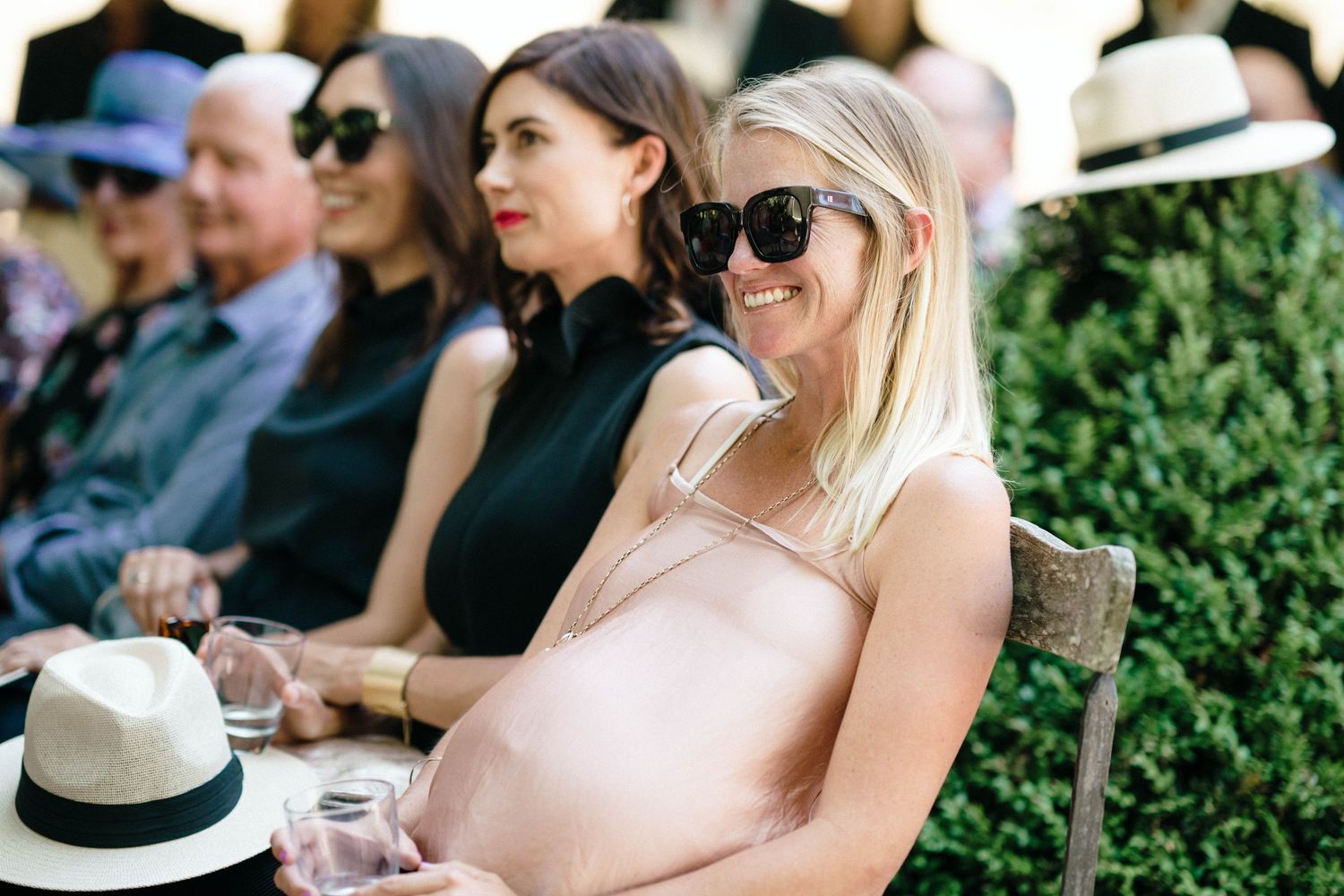 Wedding guests sit in rows at an outdoor reception with sunglasses and summer hats.