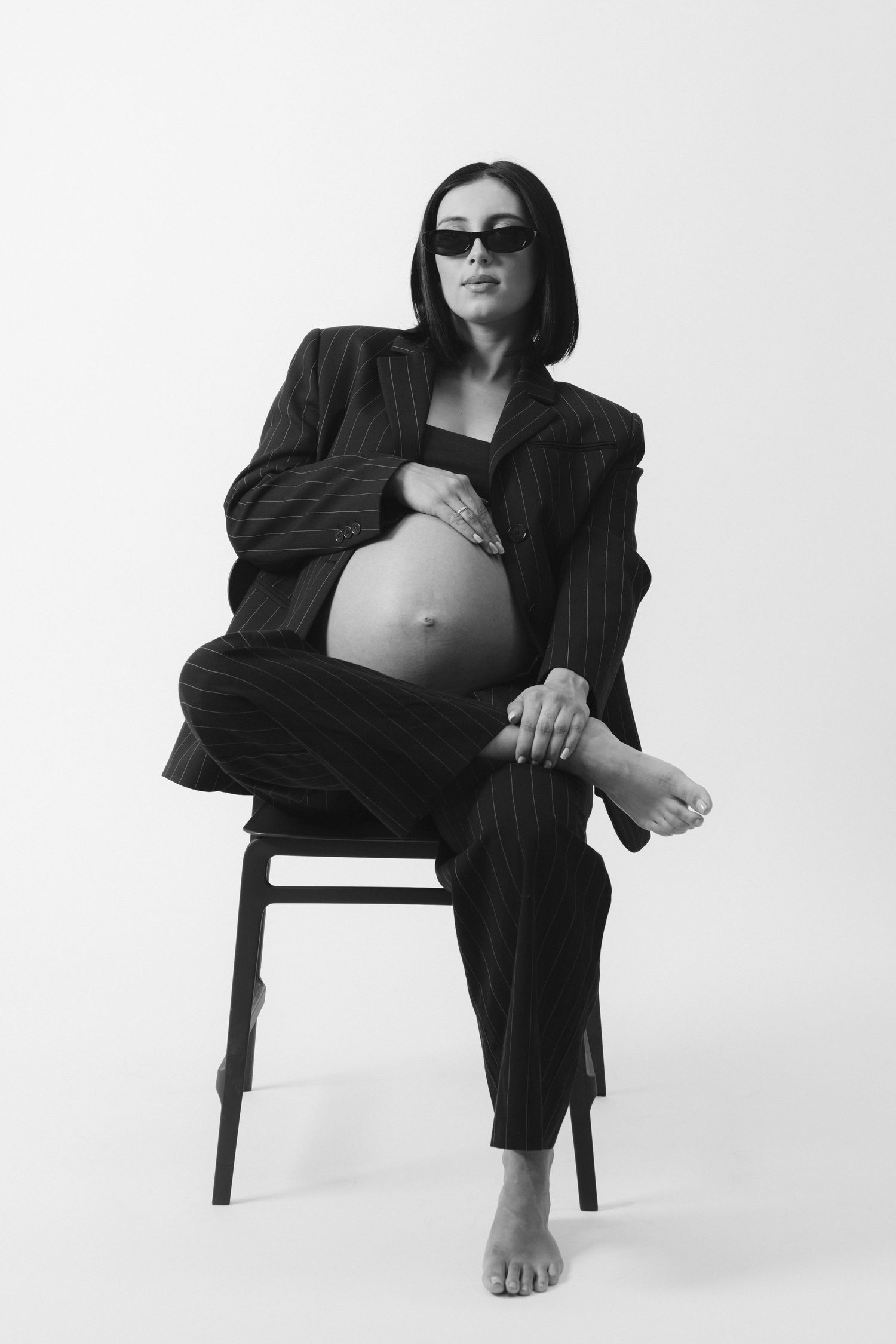 Black and white photo of a pregnant woman in a pinstripe suit and sunglasses sitting on a chair.