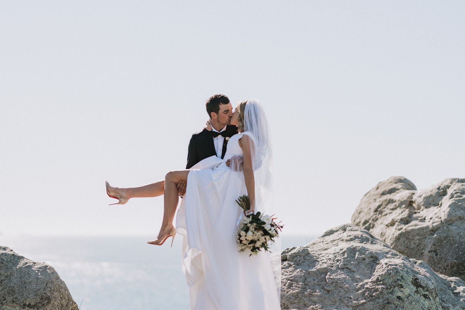 Groom in tuxedo carrying bride in white wedding dress on rocky seaside cliff during romantic outdoor ceremony.
