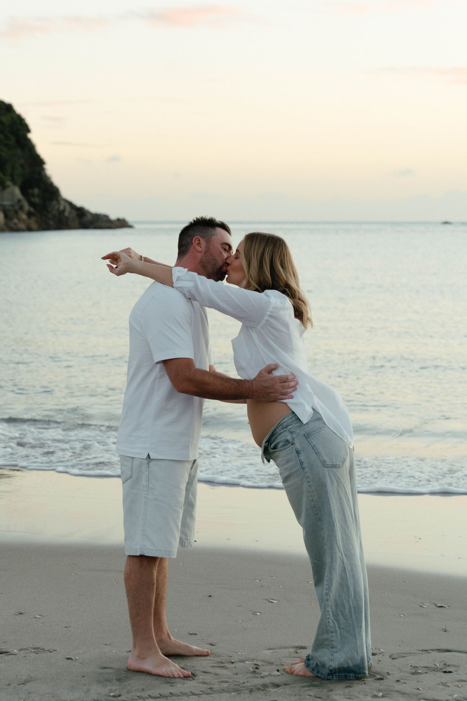 Couple kissing on a sandy beach at sunset, man in white shirt pointing while embracing pregnant woman in jeans. Bryony at Newfound.