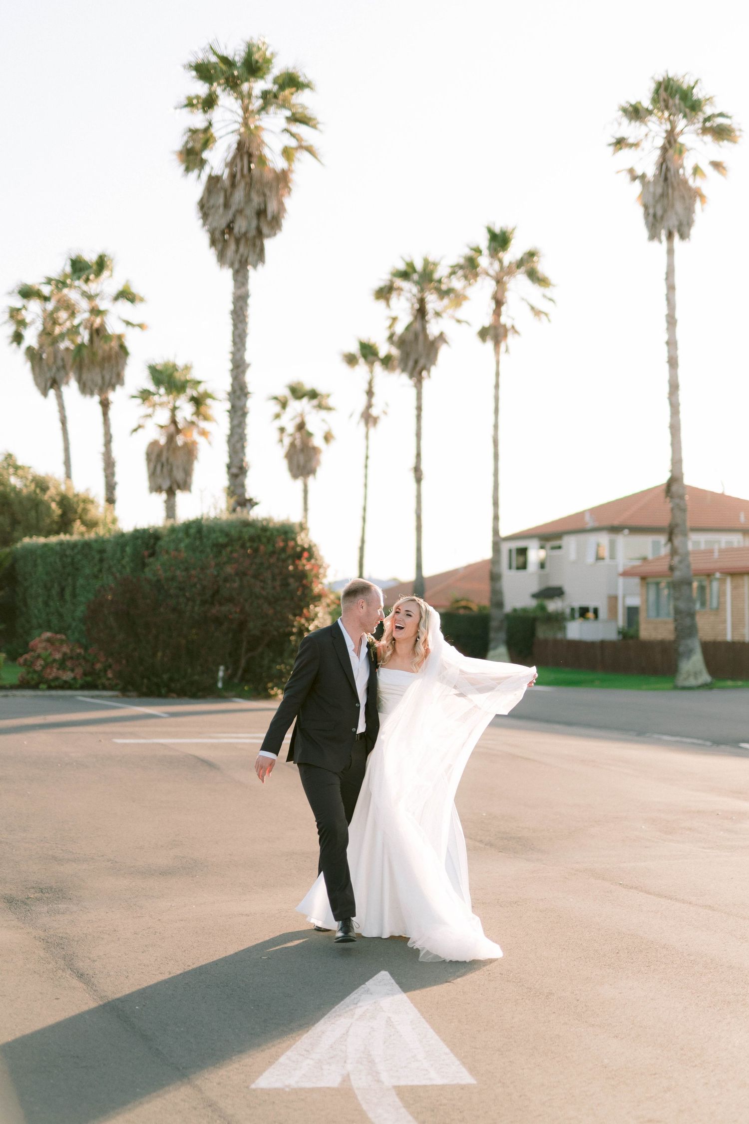 Bride and groom walking on a sunlit street lined with tall palm trees, bride's white gown flowing in the breeze.