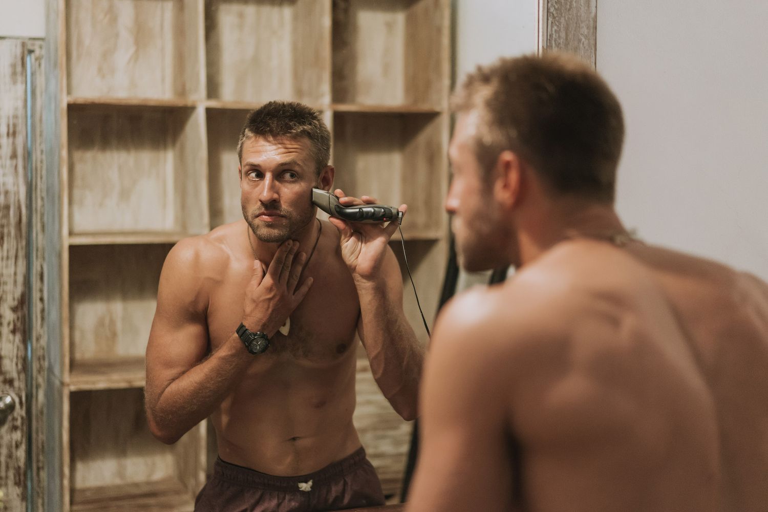 A shirtless person shaving in front of a bathroom mirror with wooden shelving in the background.