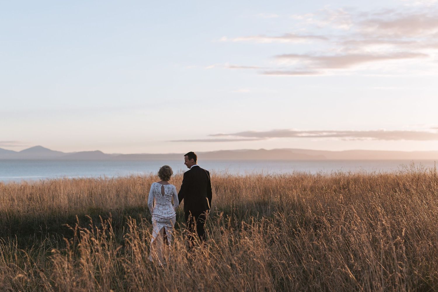 Two silhouetted figures stand in tall golden grass overlooking mountains during sunset.