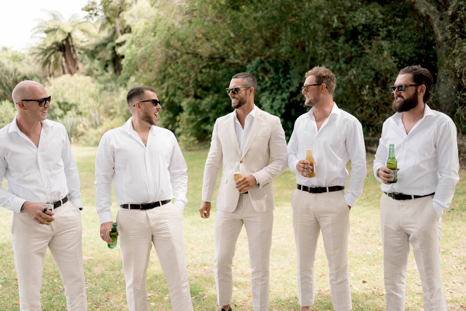 A lineup of groomsmen in crisp white shirts and slacks share laughs and smiles during an outdoor wedding photo session.