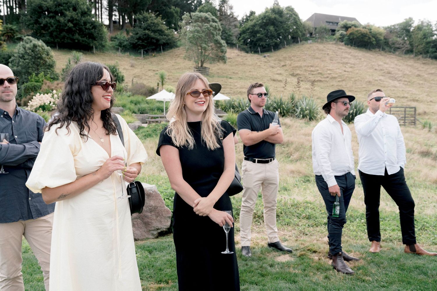 A casual outdoor gathering of people standing on a grassy hillside on a sunny day.