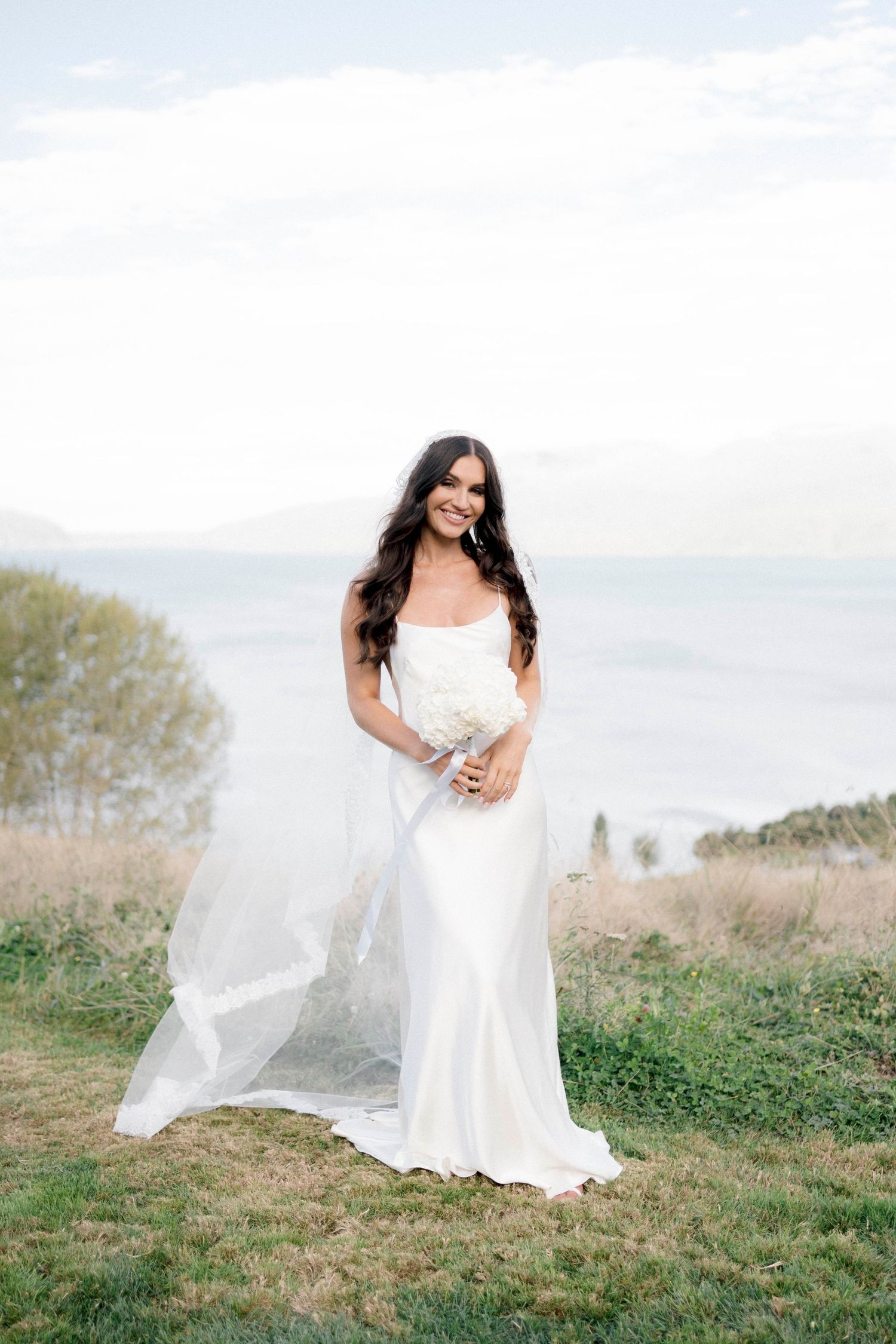 A bride in white dress twirling in a grassy field under cloudy skies.