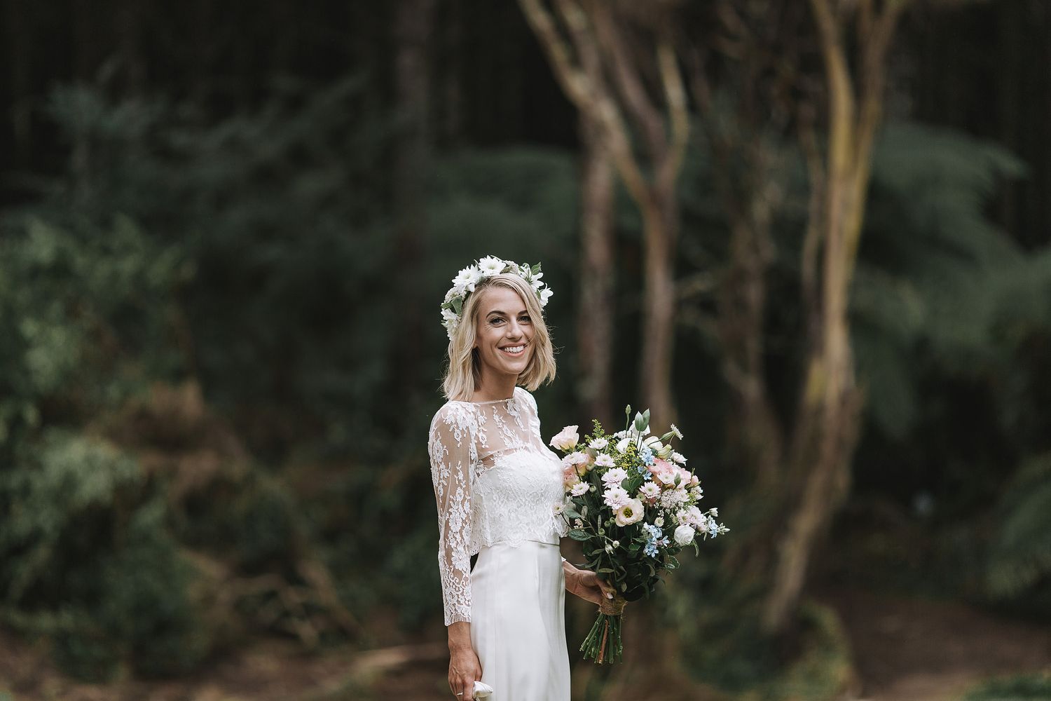 A bride in a long-sleeved lace gown and floral crown holds a white and green bouquet in a forested setting.