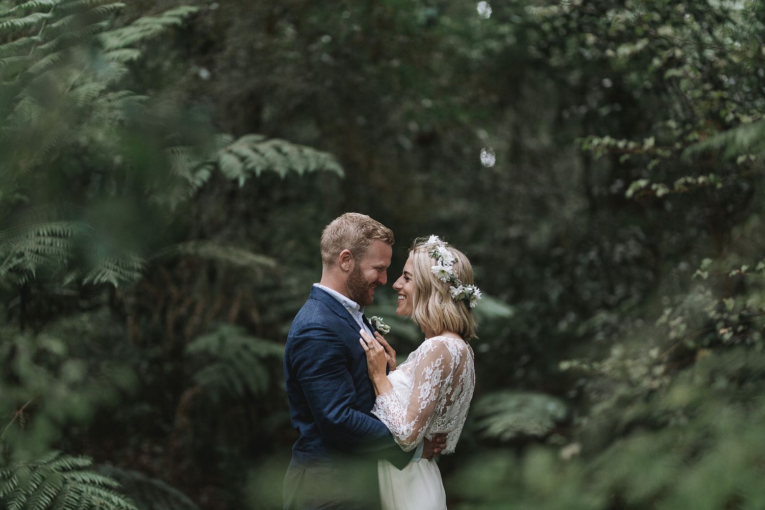 Two people share a romantic kiss outdoors surrounded by lush green foliage at a wedding celebration.