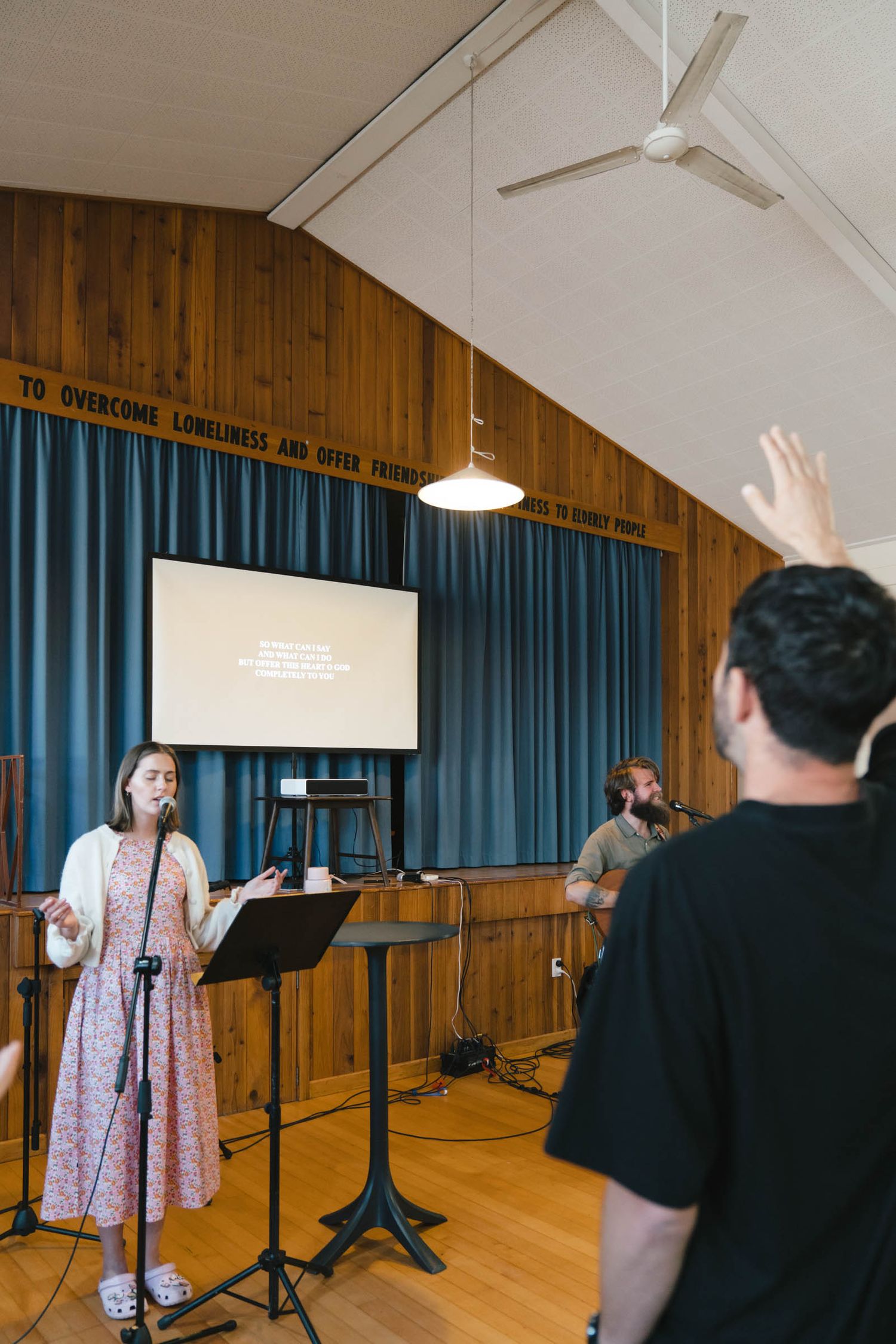 Performance space with wooden walls and blue curtains featuring musicians and projection screen.
