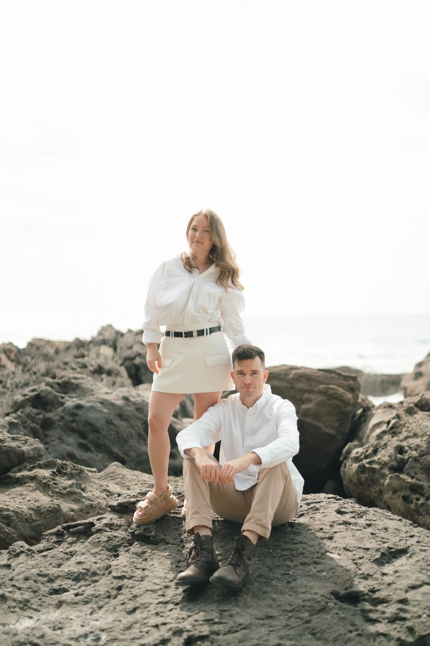 A couple sits together on rocky coastal terrain wearing coordinated white and beige outfits at sunset.
