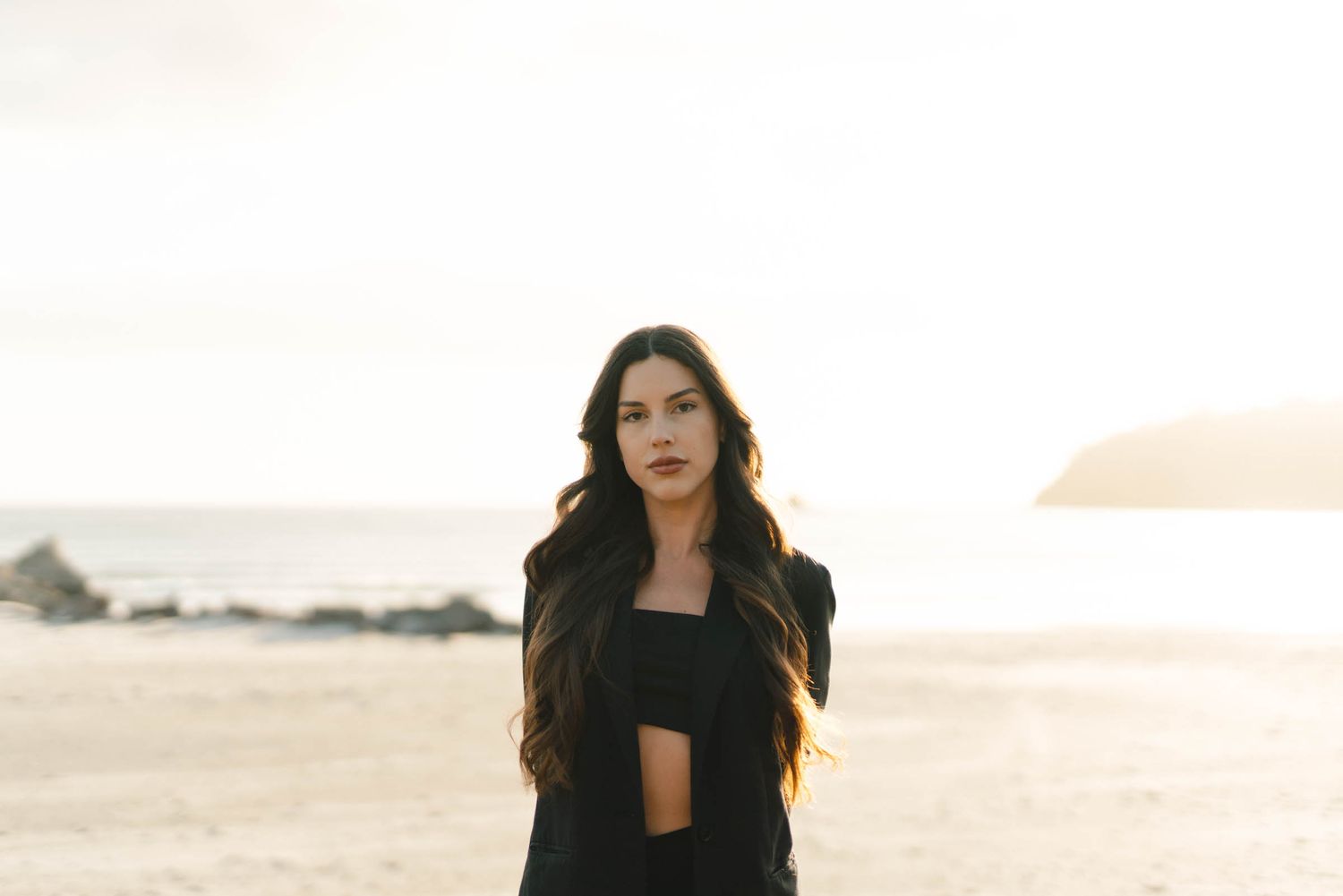 Portrait of long-haired figure in black attire against golden sunset landscape.