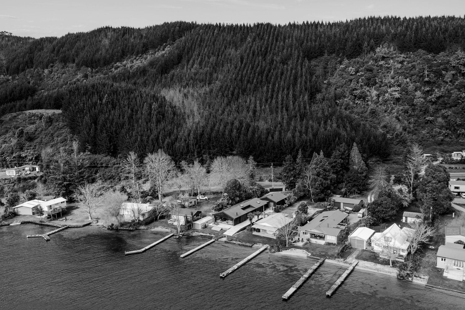 Aerial black and white view of waterfront homes with long wooden docks extending into a bay beside forested hillsides.