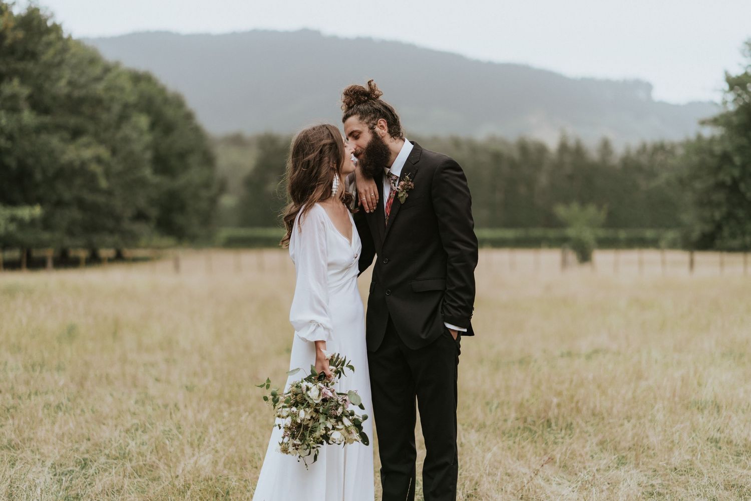 A romantic couple in wedding attire share an intimate moment in a field with mountain landscape in the background.