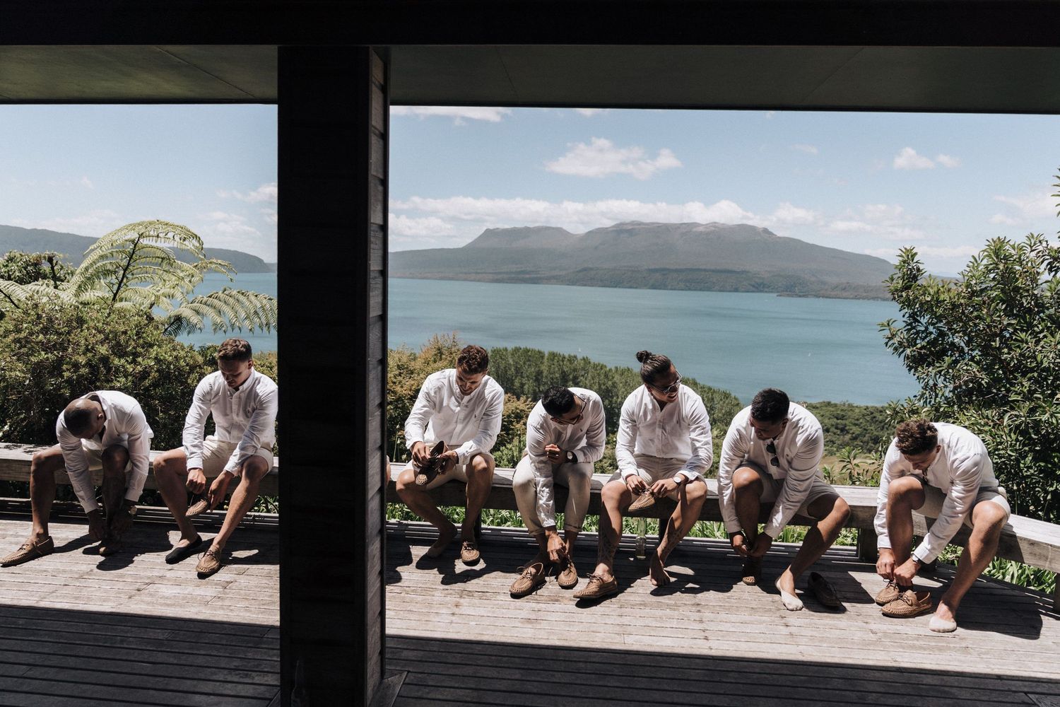 Groomsmen in white shirts sit together on a deck overlooking a scenic mountain lake view on a sunny day.