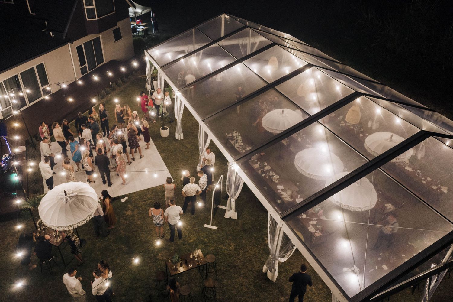 Night view of an illuminated clear tent wedding reception with guests mingling on the lawn.