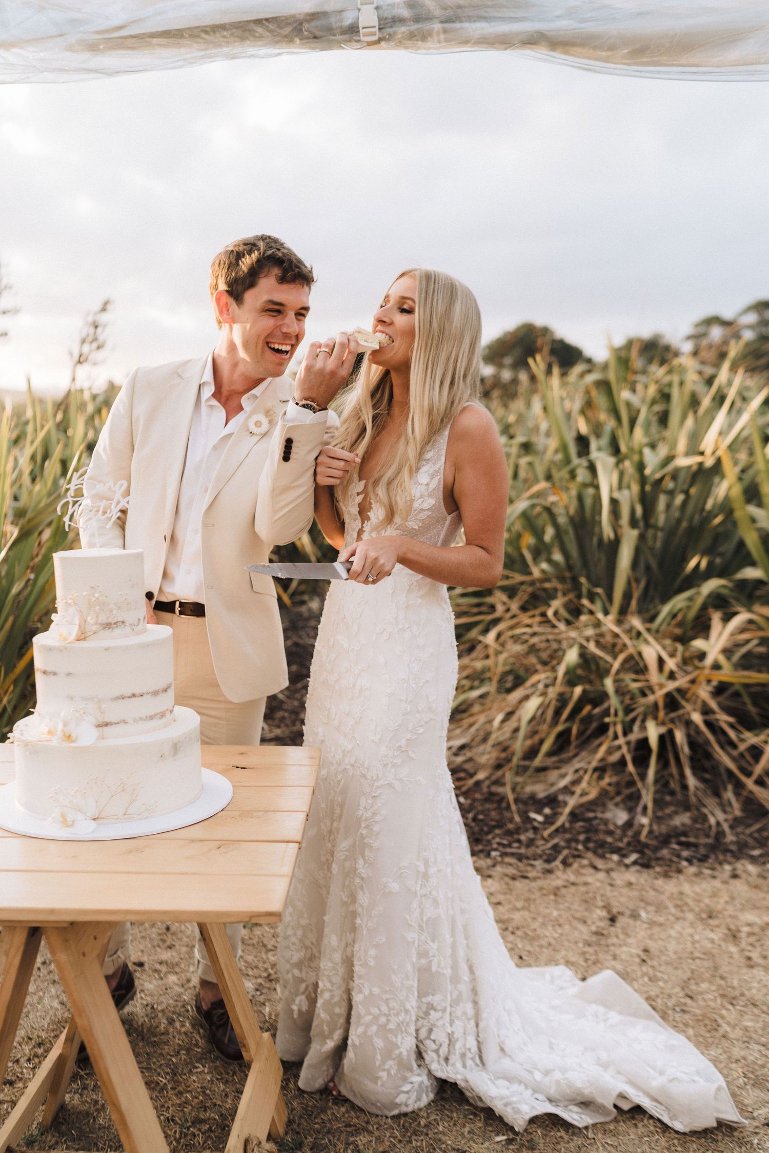 Newlywed couple sharing wedding cake at an outdoor reception with tall grass and plants in the background.