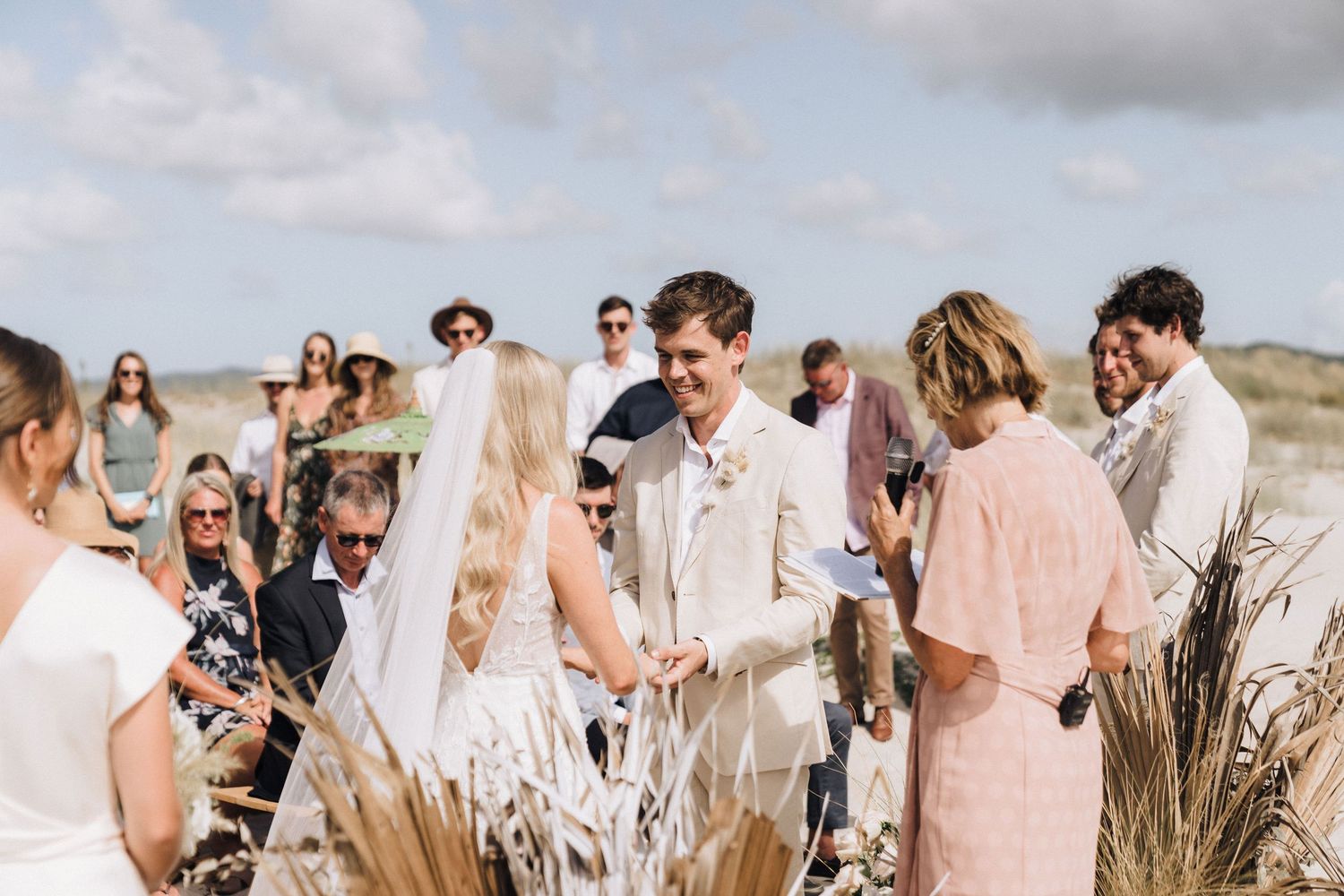 A joyful outdoor beach wedding ceremony with guests gathered around the couple in light-colored attire and pampas grass decor.