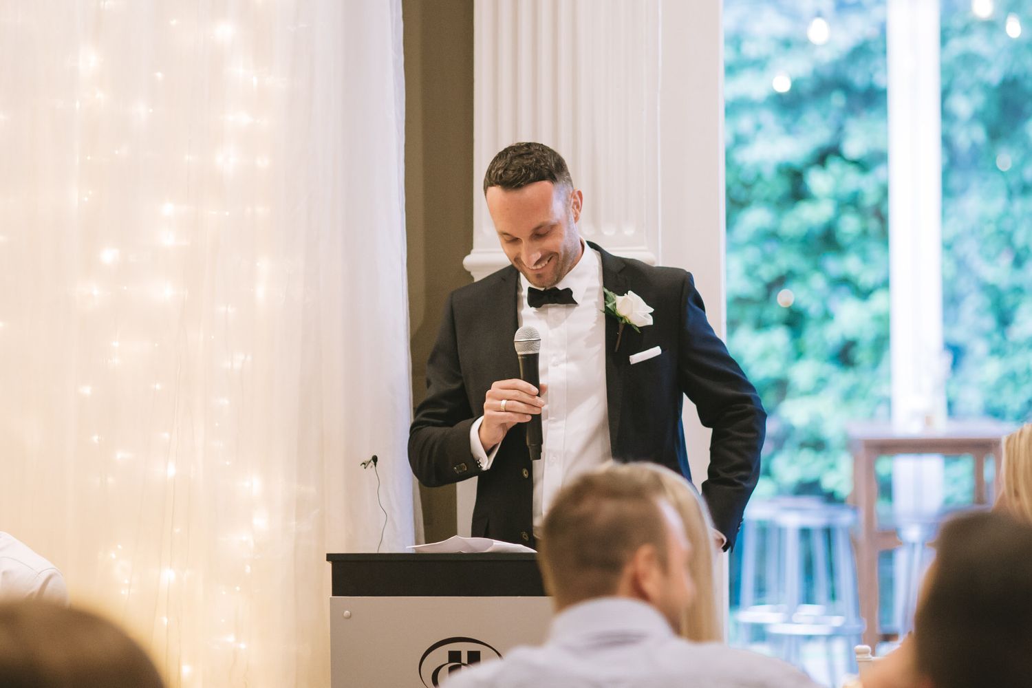 Someone in a black tuxedo delivers a speech at an elegant wedding reception while standing at a podium.