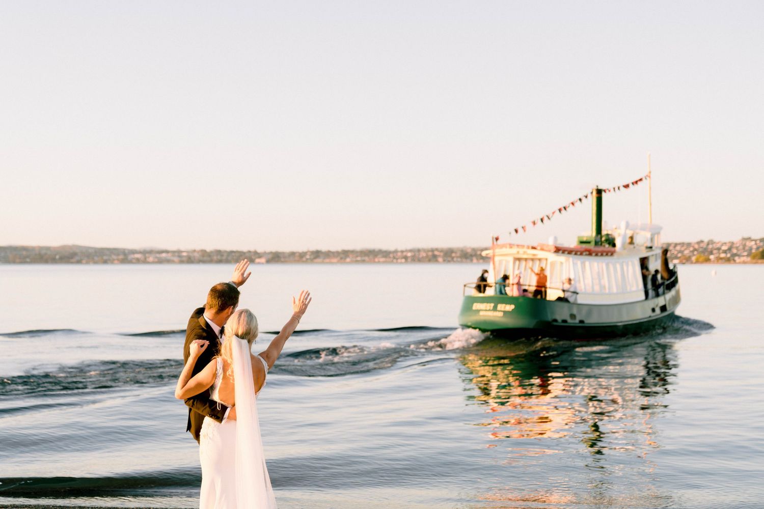 Person in white dress waves to a boat departing on calm waters with string lights at sunset.