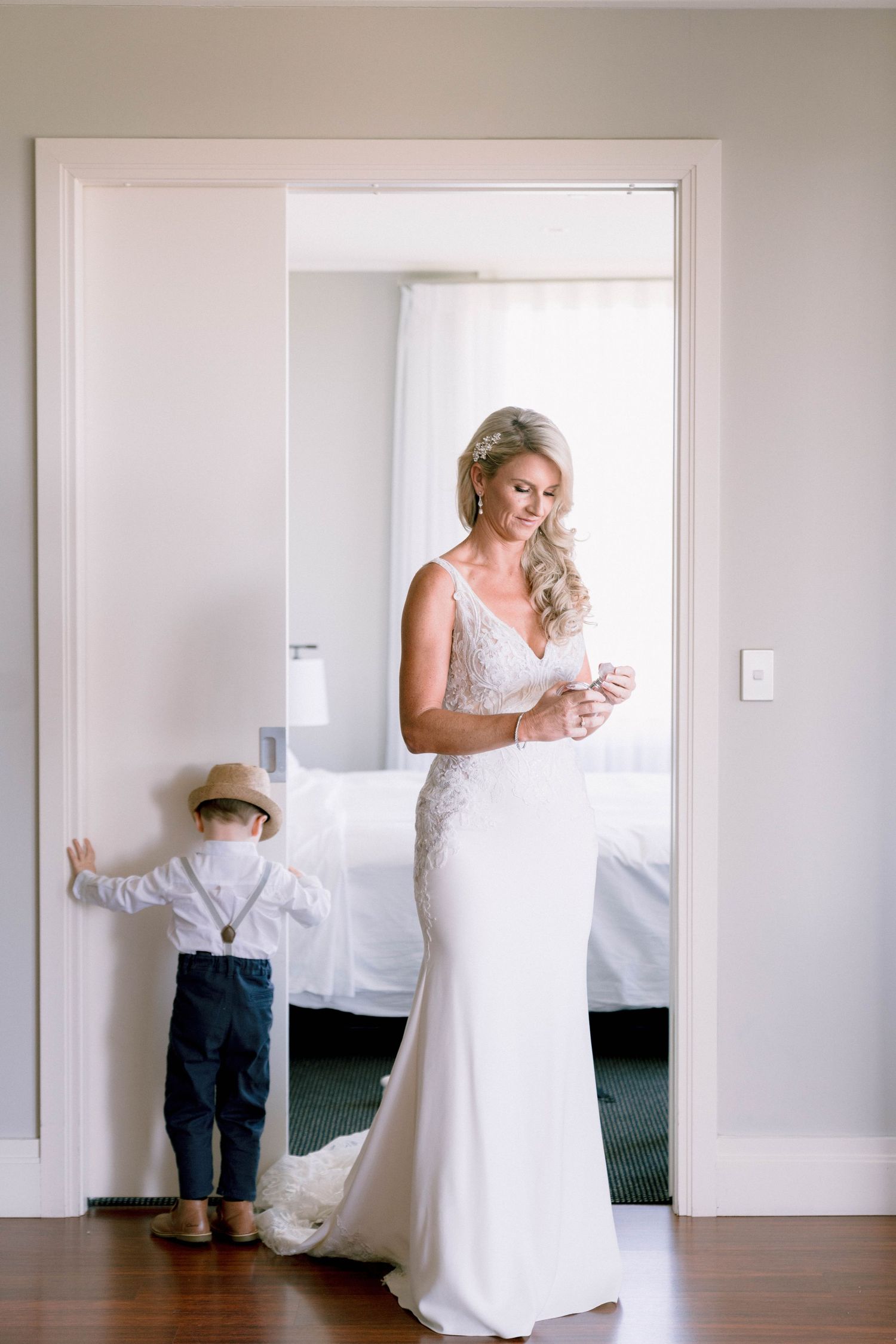 A sweet moment shows a bride in white lace gown standing in a doorway while a small boy peeks around the corner.