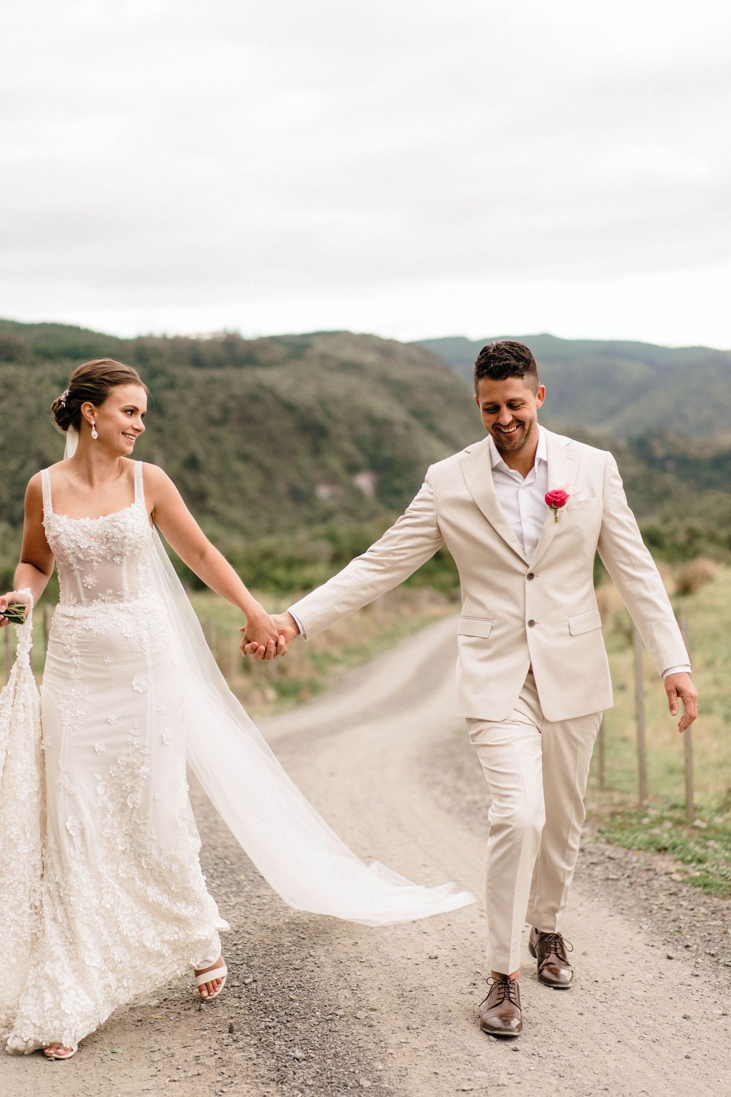 Newlyweds walking hand in hand on a dirt road with mountain views.
