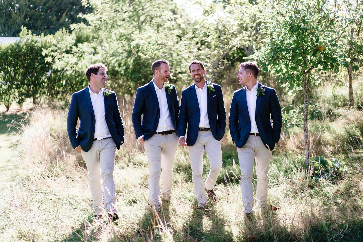 Four groomsmen in matching navy blazers and white pants pose together in sunny garden setting.