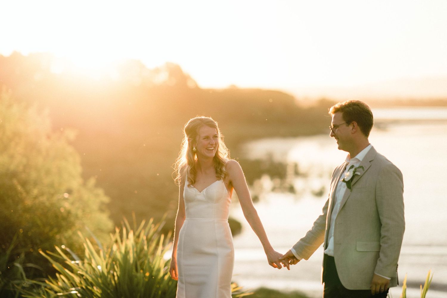 Couple holding hands at sunset by the water in formal wedding attire.