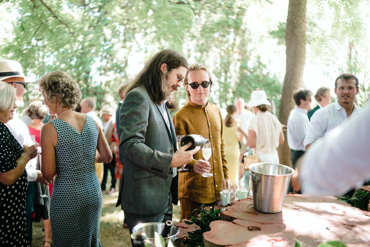 Guests mingling at an outdoor party bar area under shaded trees.