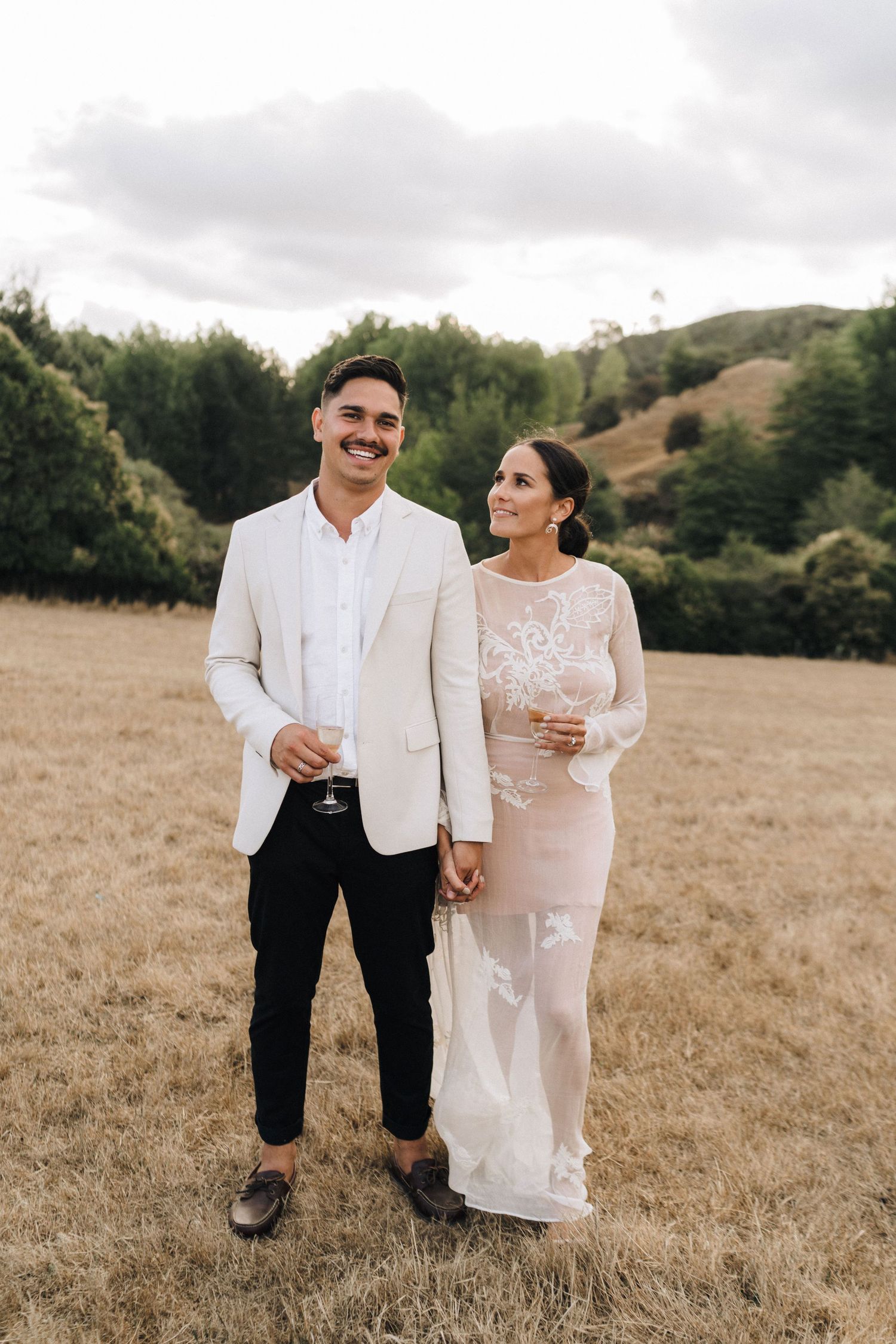 A couple poses together in formal wedding attire in a golden field with hills in the background.