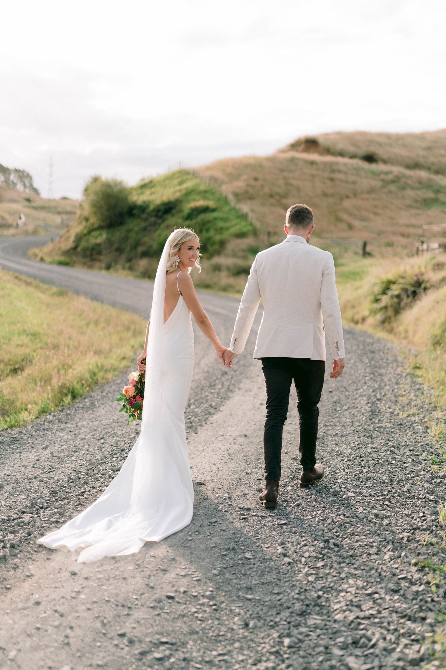 Newlyweds walking hand in hand on a gravel path through rolling hills.