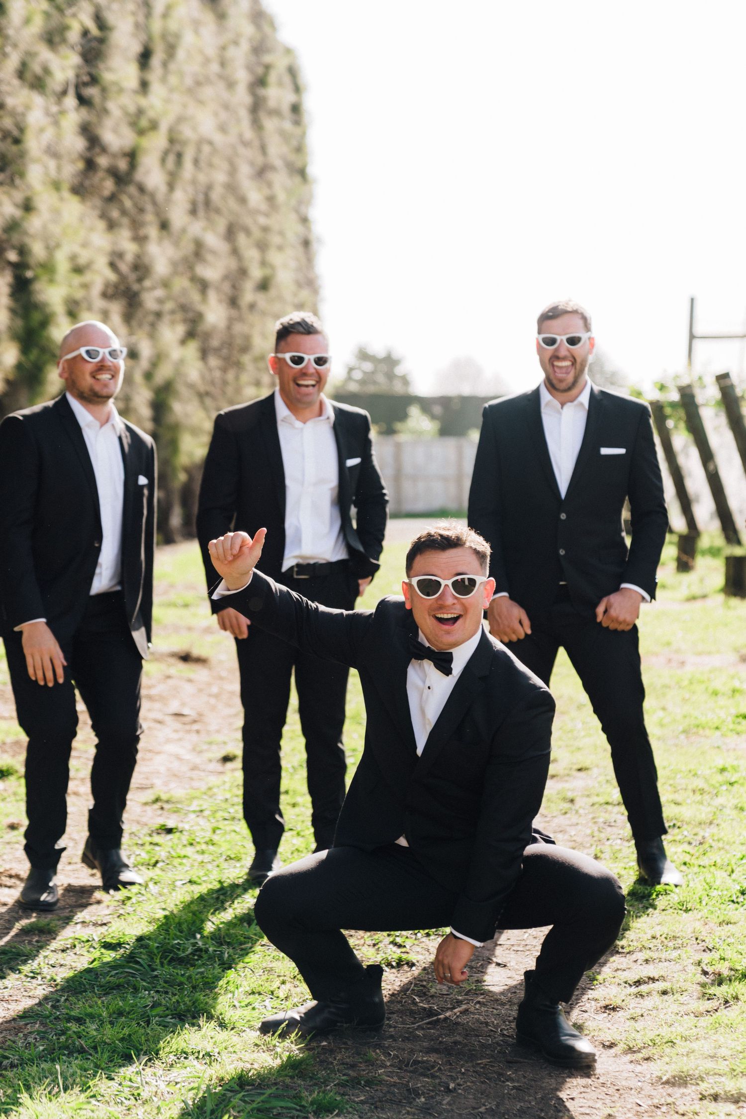 Groomsmen in black tuxedos and sunglasses pose playfully outdoors.