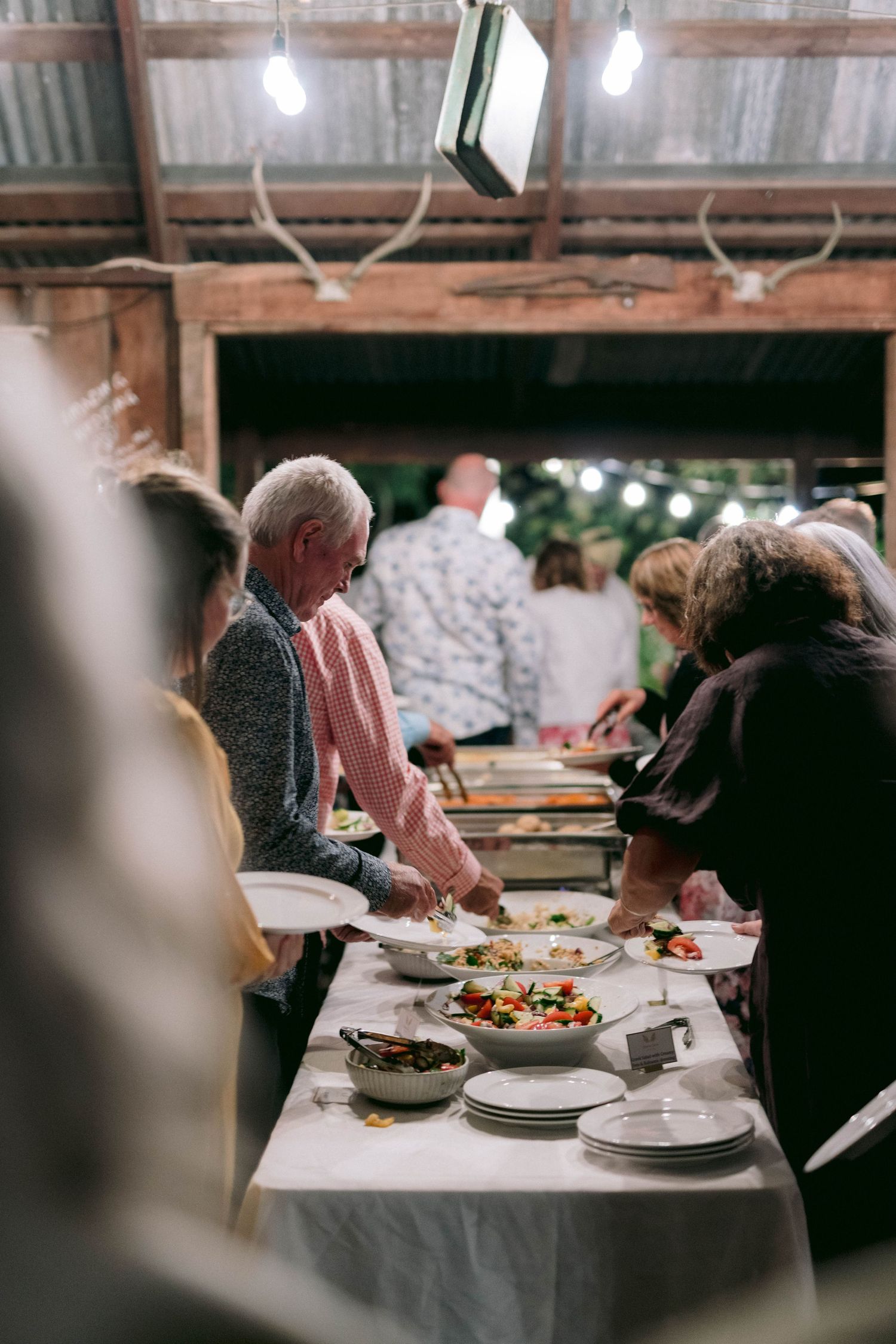 Guests serving themselves at a wedding buffet table under rustic lighting.