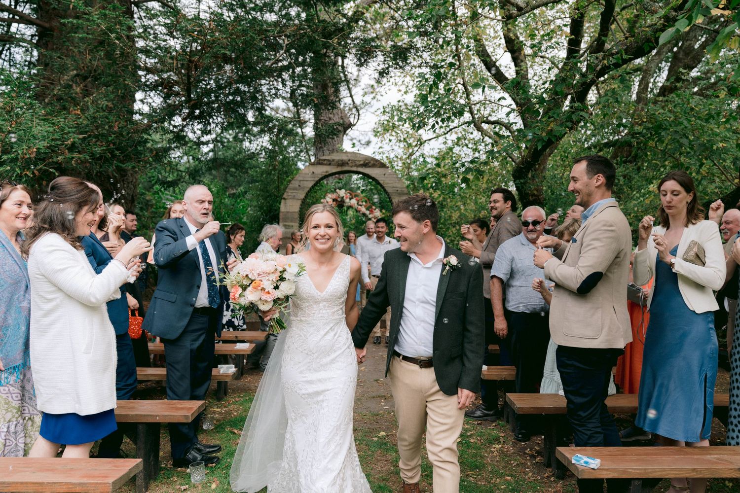 Wedding guests applaud as newlyweds walk down the aisle at an outdoor ceremony.