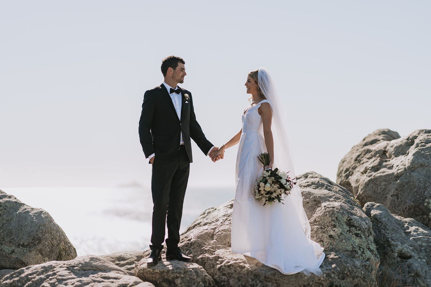 Two people stand together on dramatic rocky cliffs overlooking a scenic mountain landscape on their wedding day.