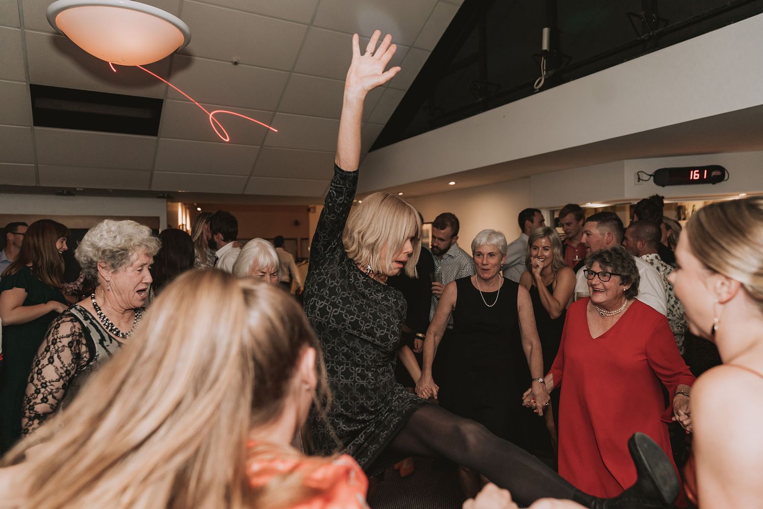 People dancing and socializing at an indoor party event with dim lighting and ceiling tiles visible.