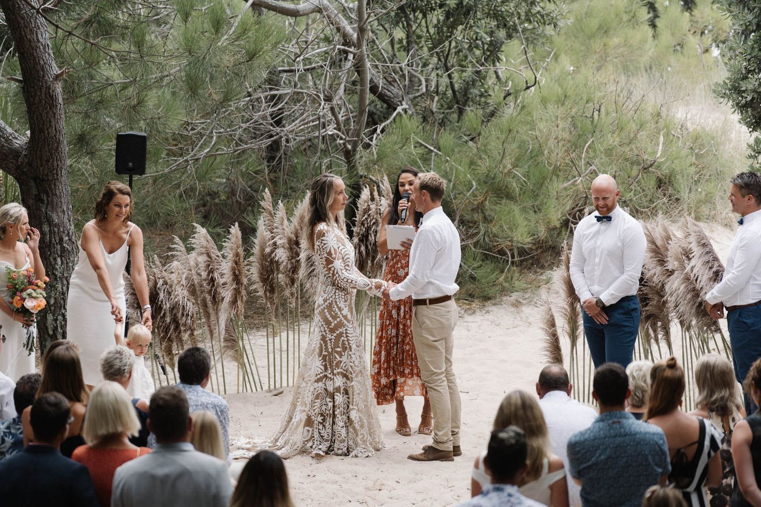 An intimate outdoor wedding ceremony takes place on a sandy path surrounded by natural greenery and pampas grass.