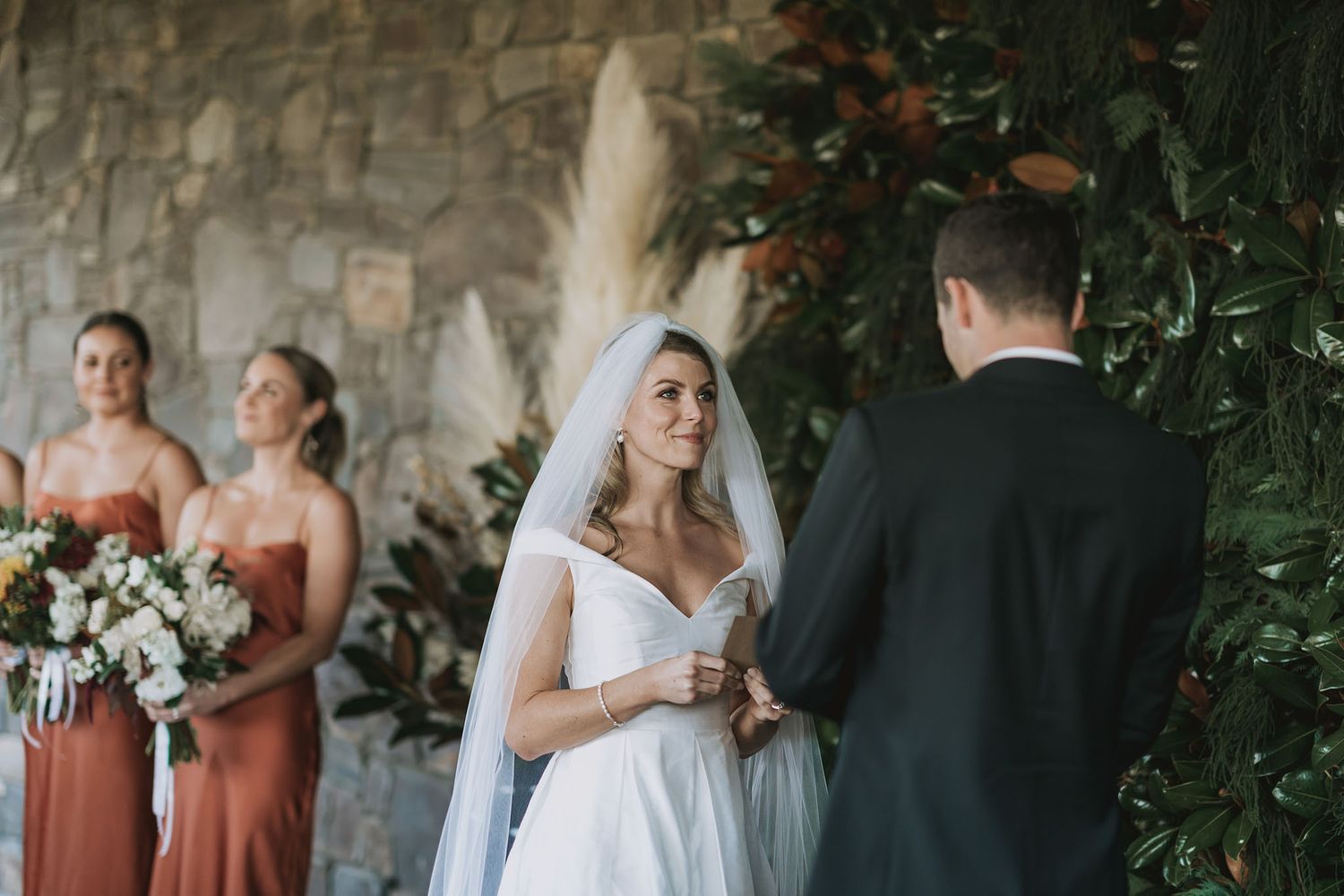 Wedding party stands in front of stone wall with lush greenery during outdoor ceremony.