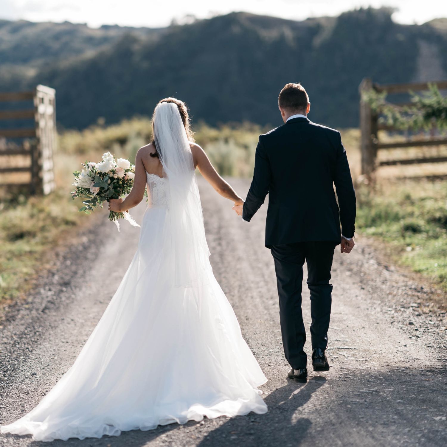 Two figures in wedding attire walk together down a rustic dirt path with rolling hills in the background.