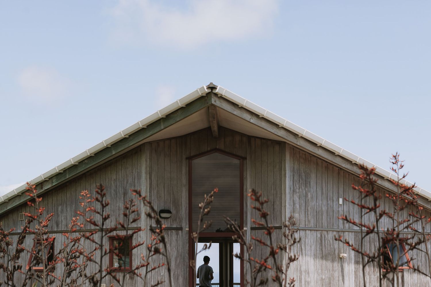 A rustic wooden barn roof peak with silhouetted figure framed in the center, surrounded by dried plant stems against blue sky.