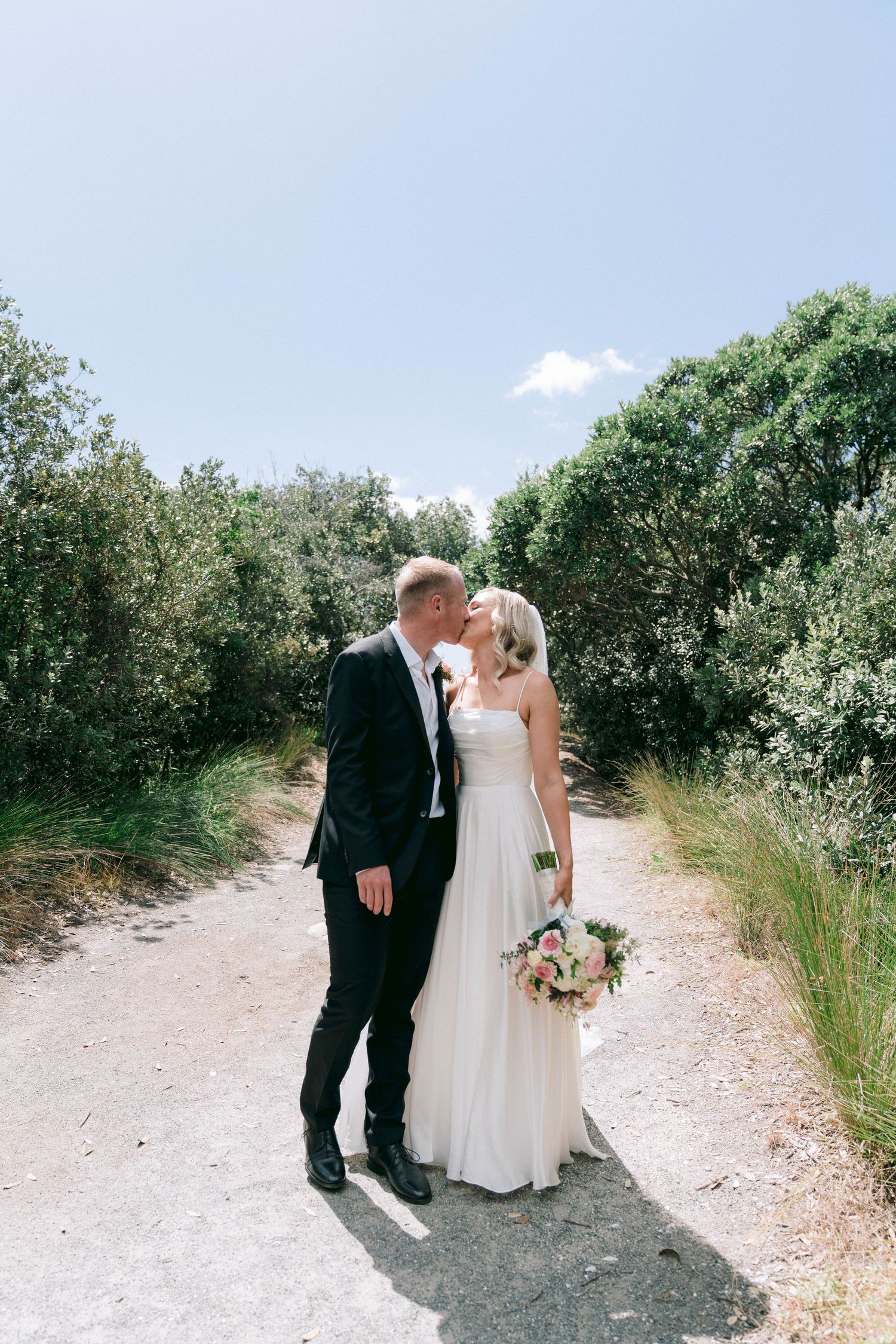 Newlyweds share a romantic moment on a sunlit dirt path surrounded by lush greenery.