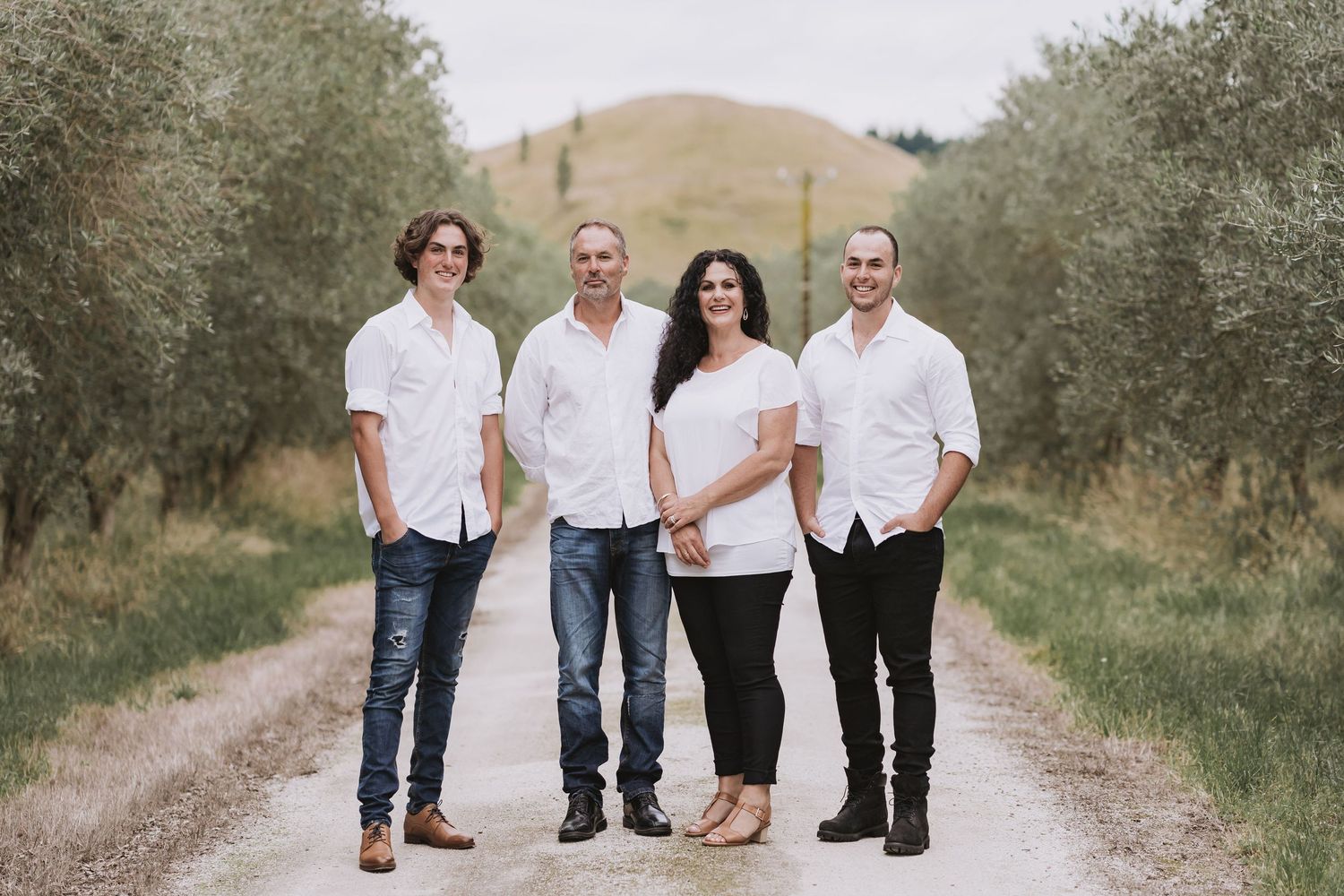 A family group poses together on a rural dirt path lined with olive trees on a sunny day.