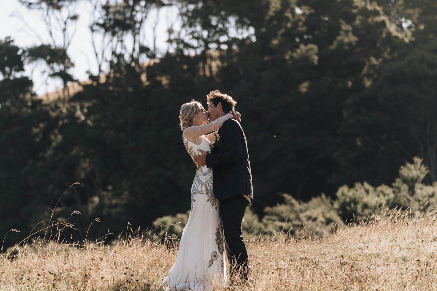 A couple holds hands and walks through a golden sun-lit meadow during their outdoor wedding photoshoot.